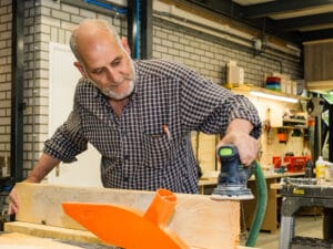 Man schuurt houten balk in werkplaats van Stichting Nieuw Ambacht tijdens houtbewerking.