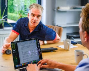 Twee mannen in gesprek tijdens een overleg bij DeJa Bouwadviseur, met een laptop en notities op tafel.