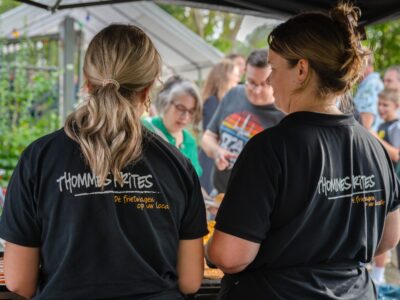 Twee medewerkers van Thommes Frites serveren snacks tijdens een levendig buurtfeest. Twee medewerkers van een frietwagen bereiden eten terwijl bezoekers wachten tijdens een zomers buurtfeest.