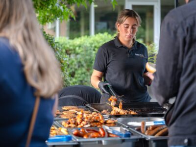 Een moment van concentratie achter de barbecue, waar de geur van vers gegrild eten en gezelligheid samenkomen. Een vrouw in zwarte bedrijfskleding bereidt gegrild vlees op een barbecue tijdens een zomers bedrijfsfeest, terwijl gasten wachten op hun maaltijd.